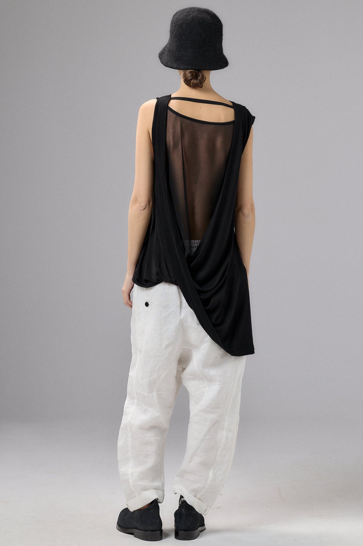 Person wearing a black sleeveless top and white pants with a black hat on a gray background