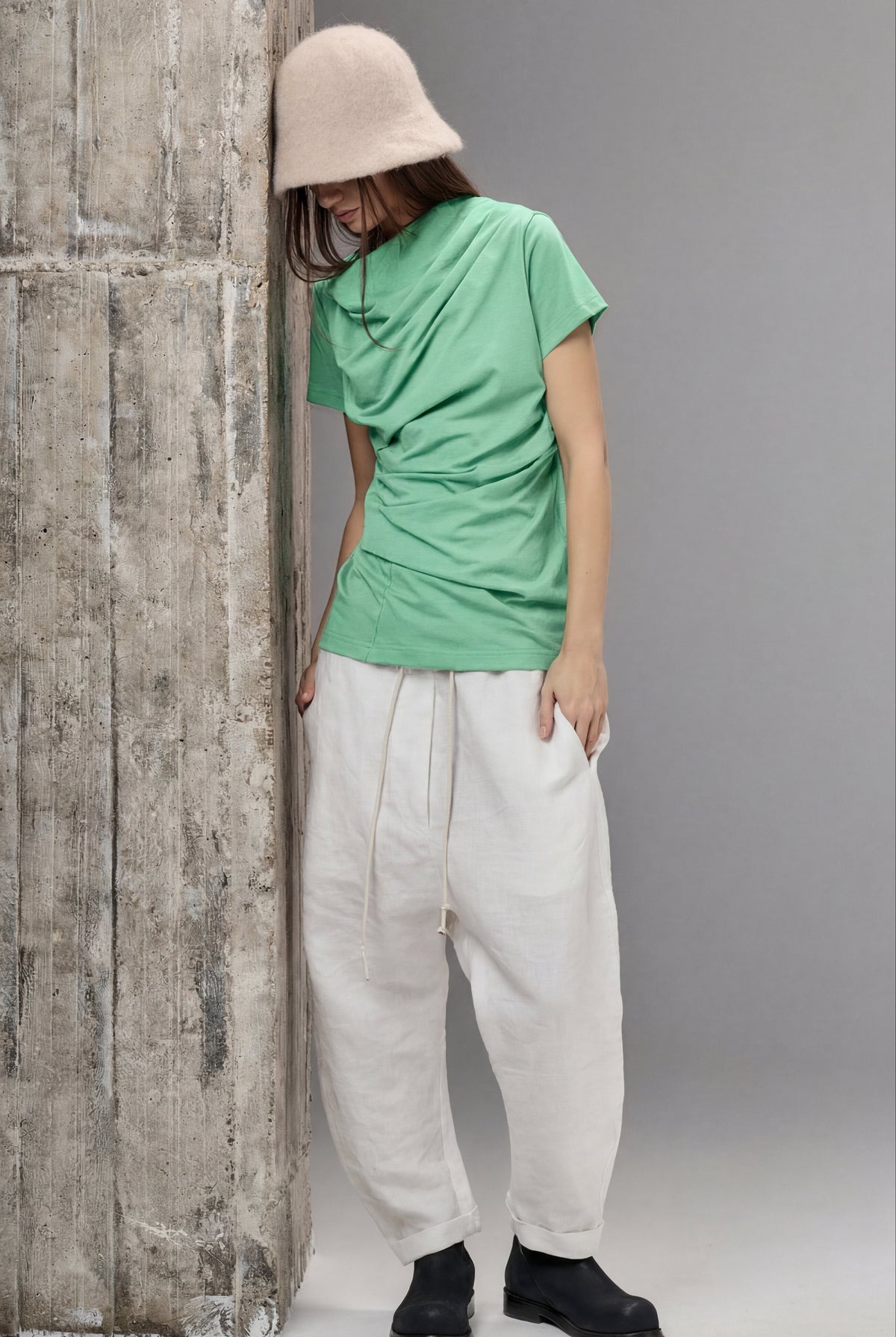 Person wearing a green shirt and white pants leaning against a textured wall.
