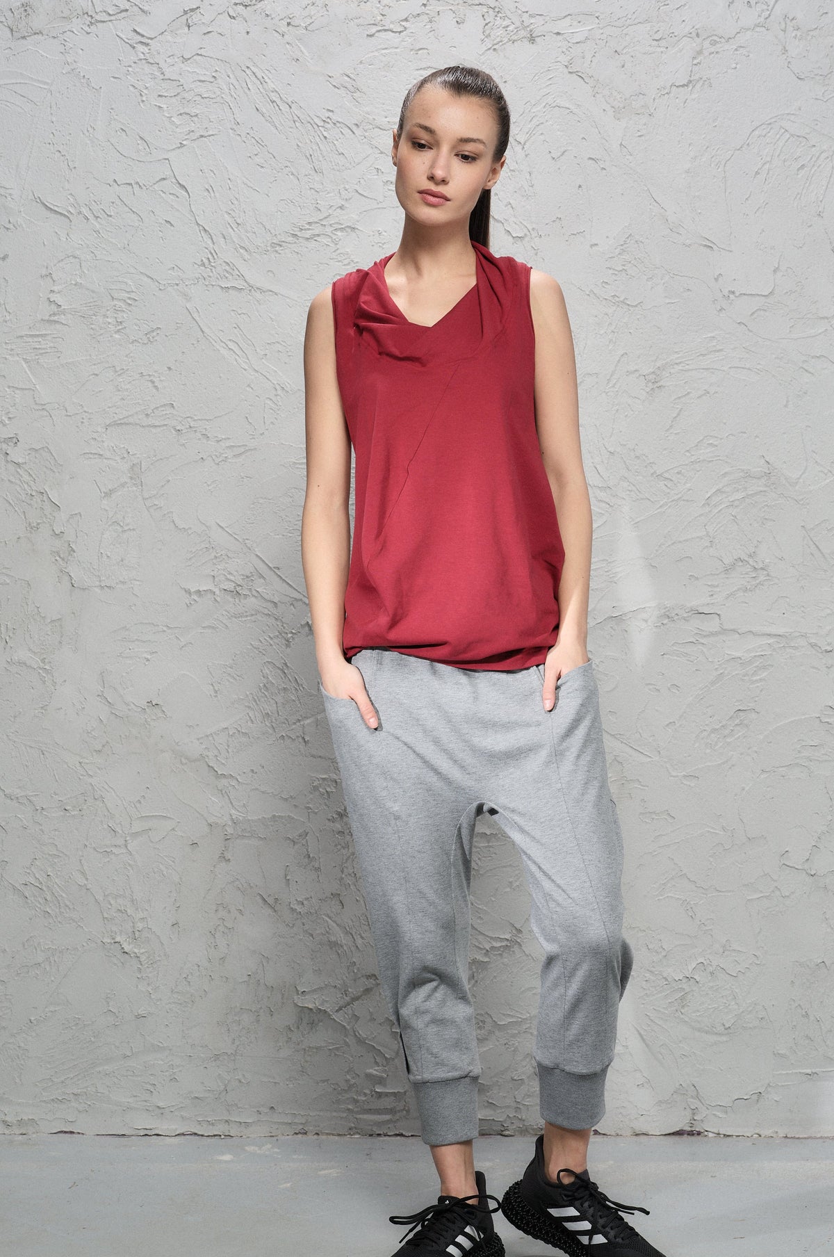 Person wearing a red sleeveless top and gray pants against a textured gray wall.