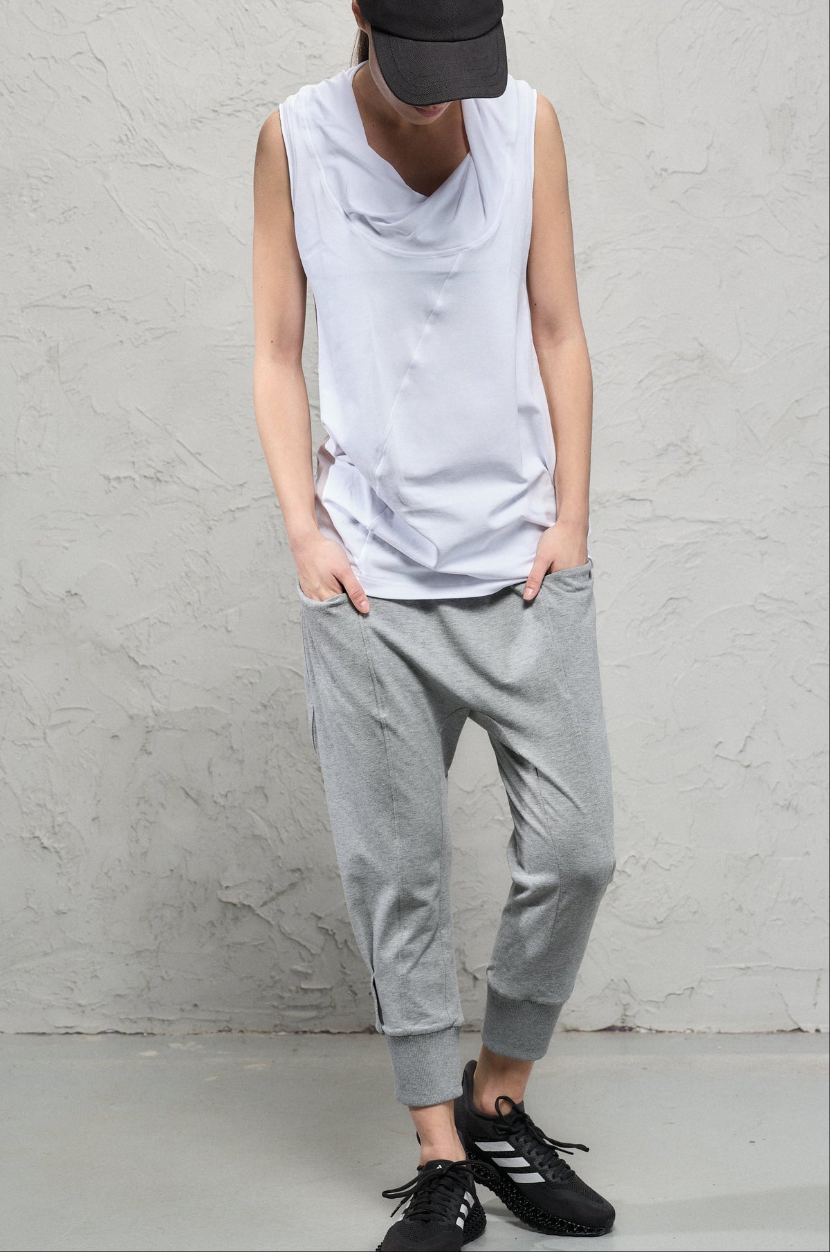 Person wearing a sleeveless gray top, gray pants, and black shoes against a plain wall.