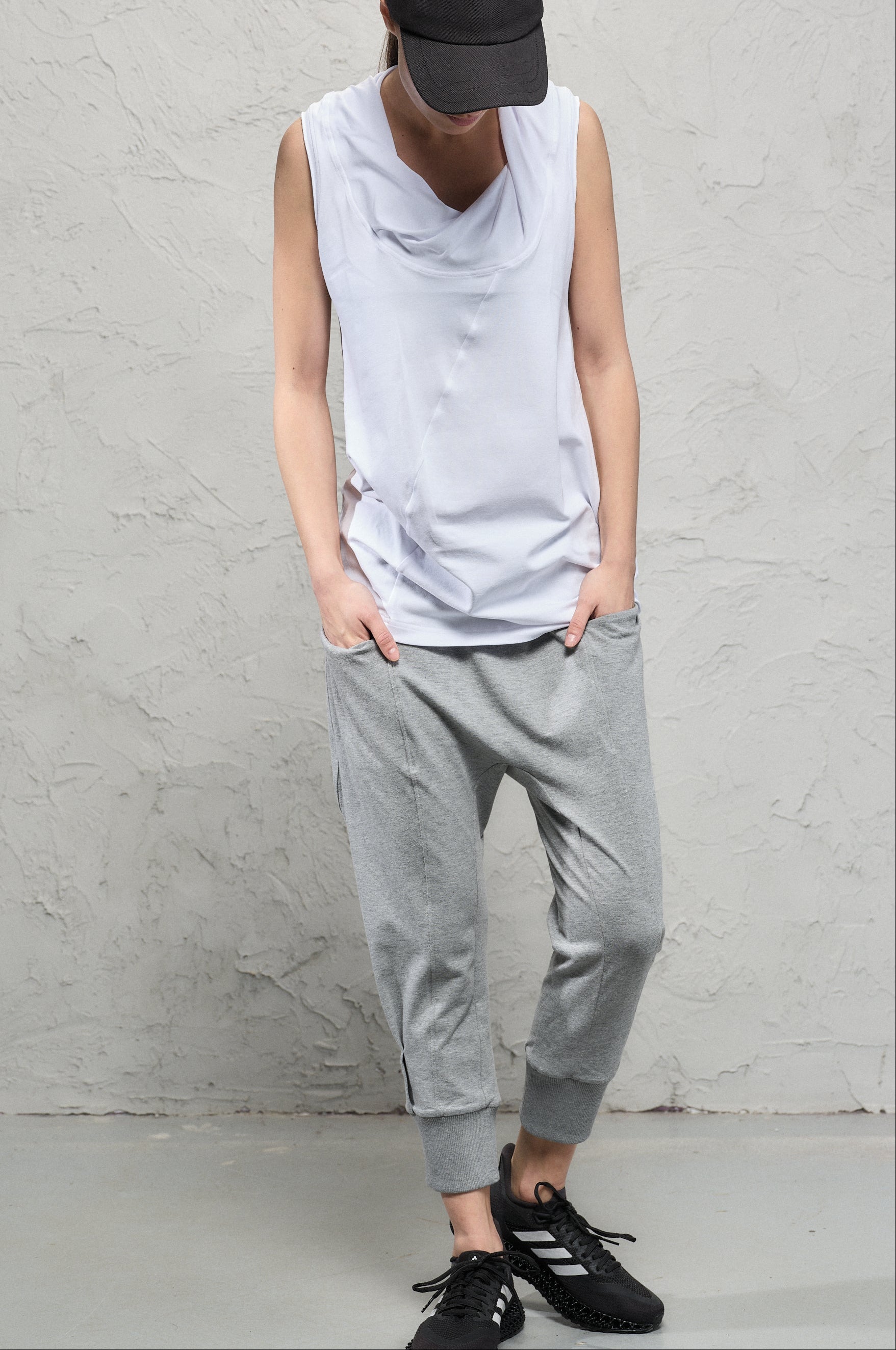 Person wearing a sleeveless gray top, gray pants, and black shoes against a plain wall.