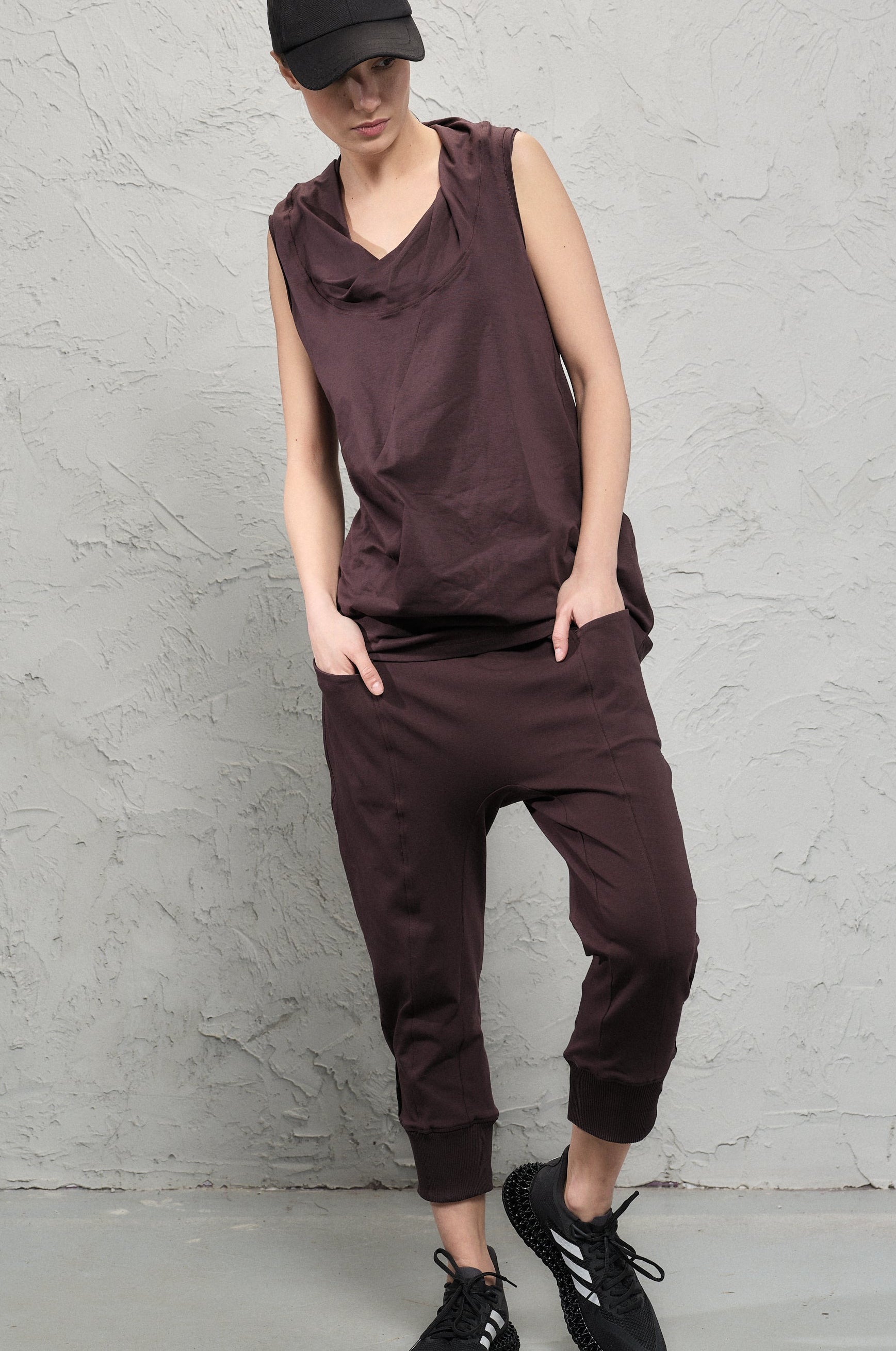 Person wearing a brown sleeveless top and pants with a black cap against a gray concrete wall.
