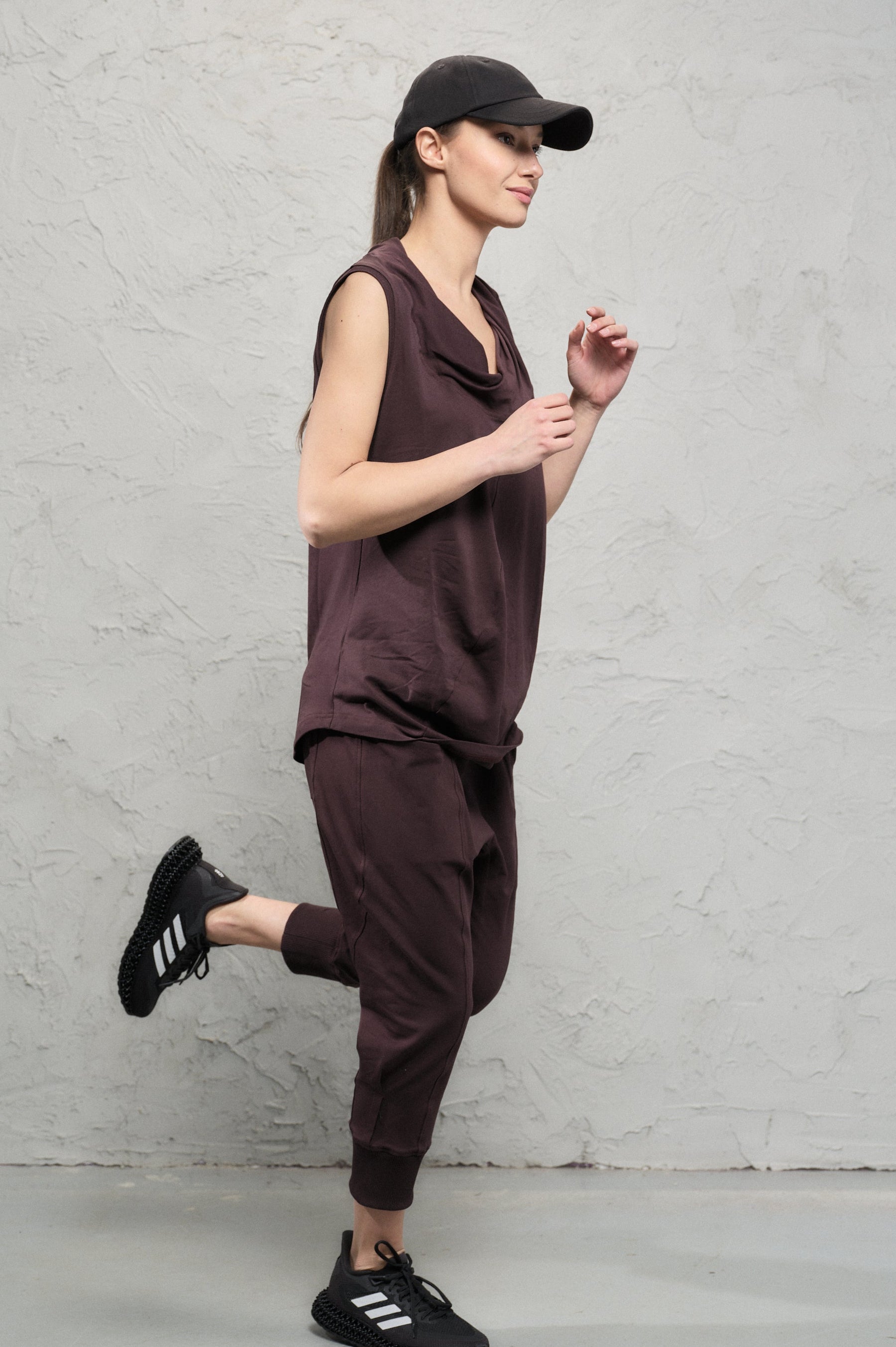 Woman in brown sleeveless top and pants with black cap and shoes against a gray background