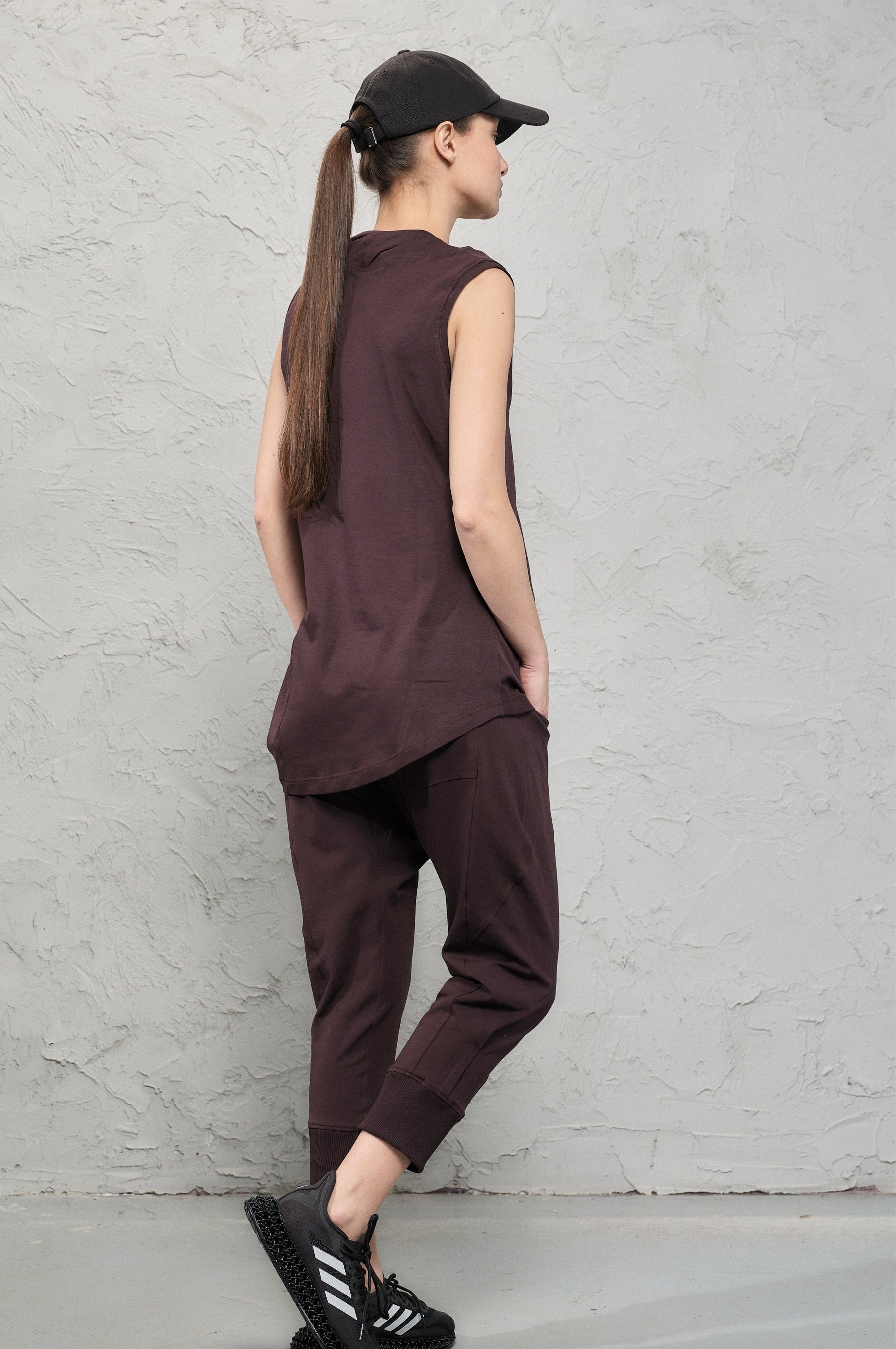 Person wearing a brown sleeveless top and pants standing against a gray wall.