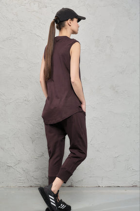 Person wearing a brown sleeveless top and pants standing against a gray wall.