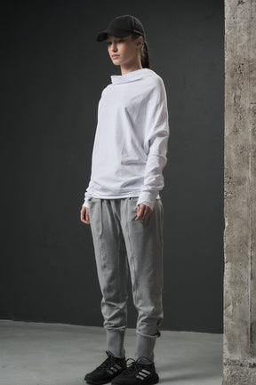 Person wearing a white hoodie, gray pants, and black shoes against a dark wall.