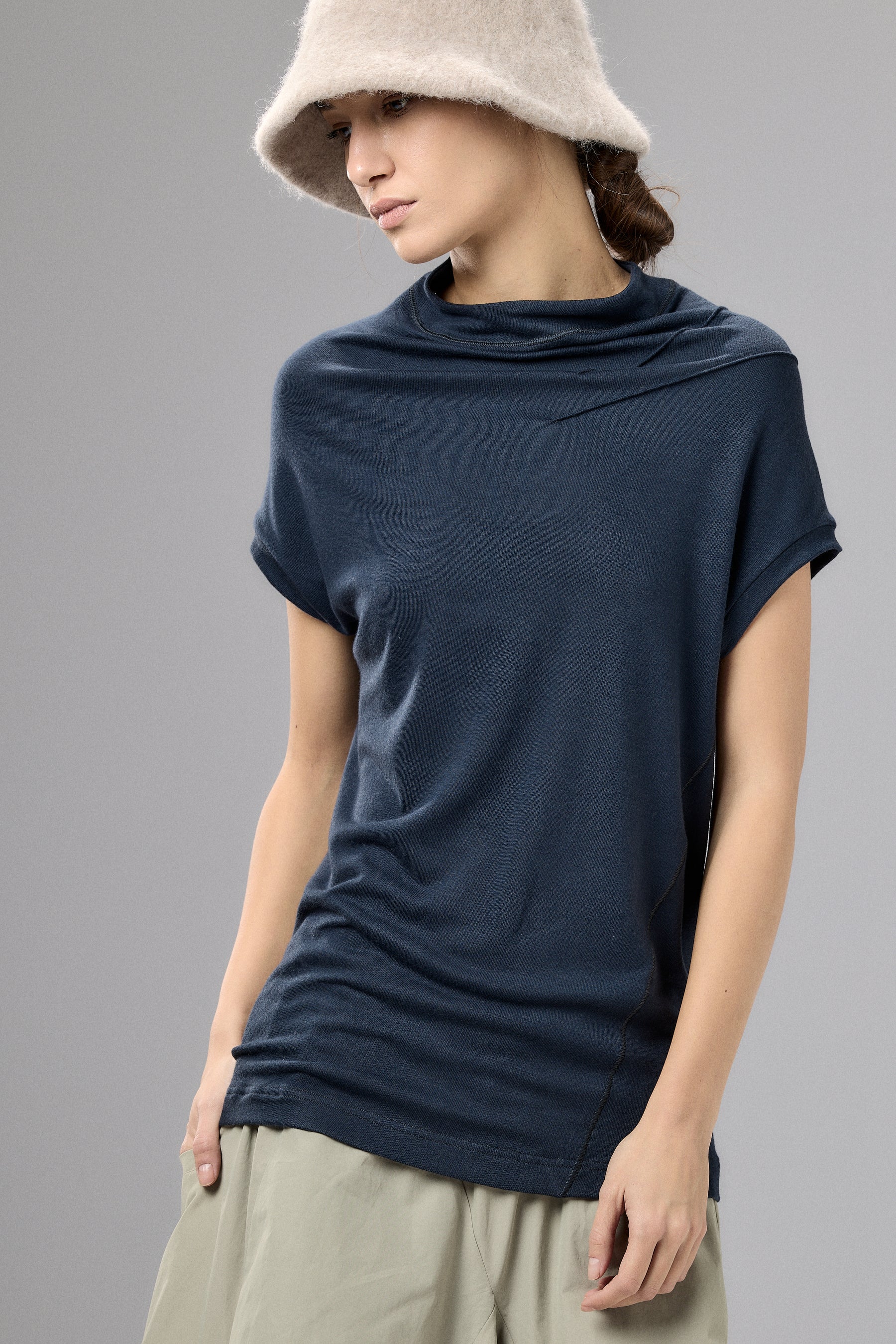 Woman wearing a navy blue cowl-neck top and beige hat on a gray background