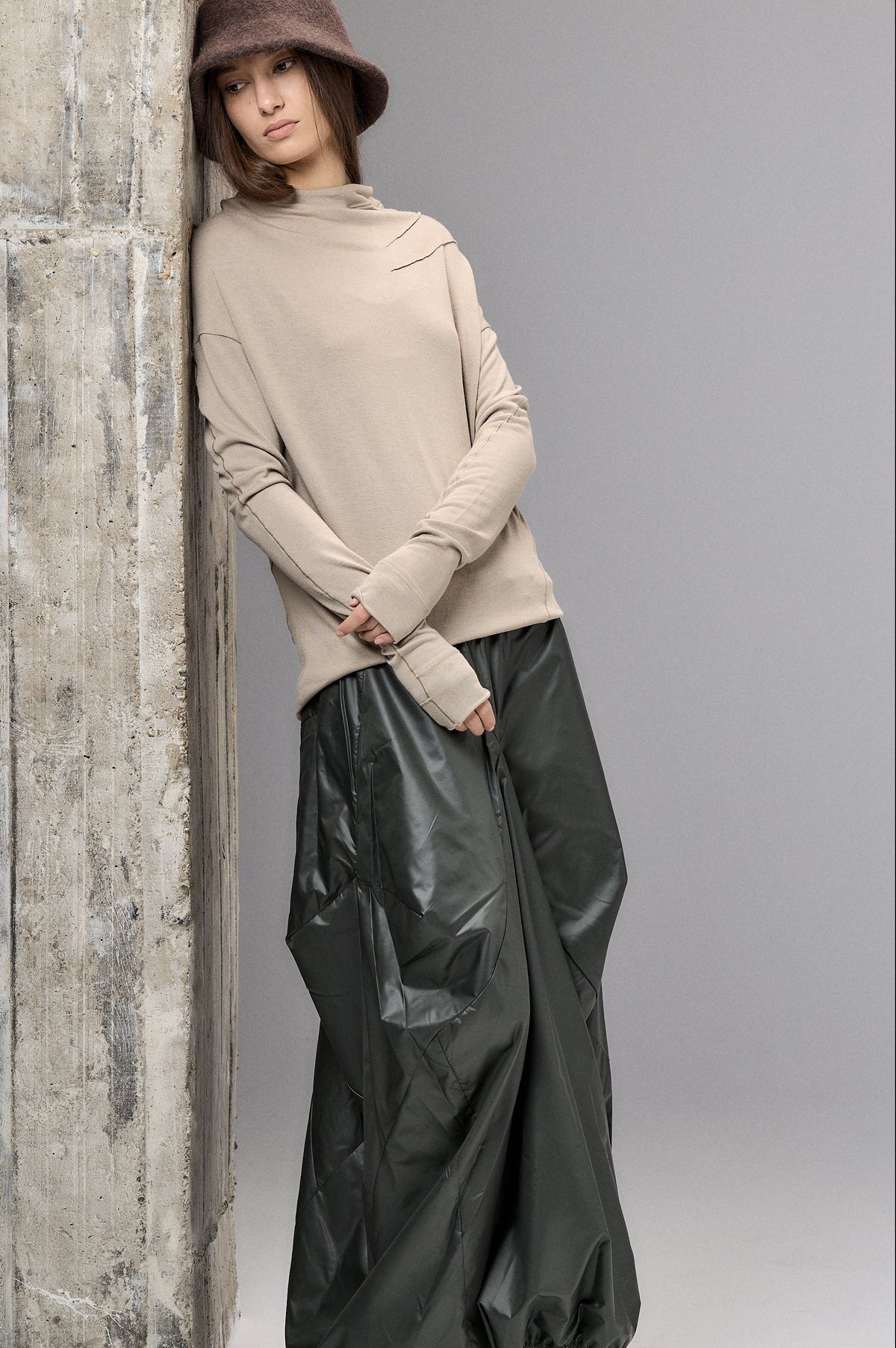 Woman wearing a beige sweater and black pants leaning against a textured wall.