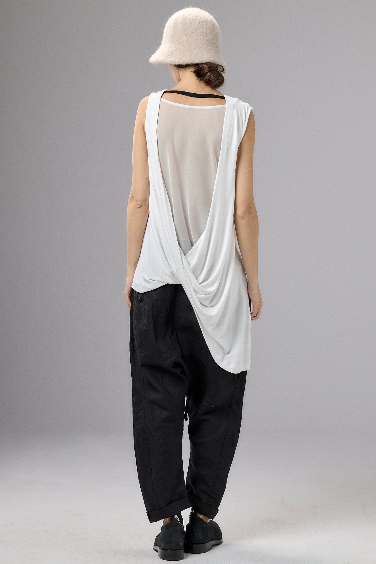 Person wearing a white sleeveless top and black pants with a beige hat on a gray background