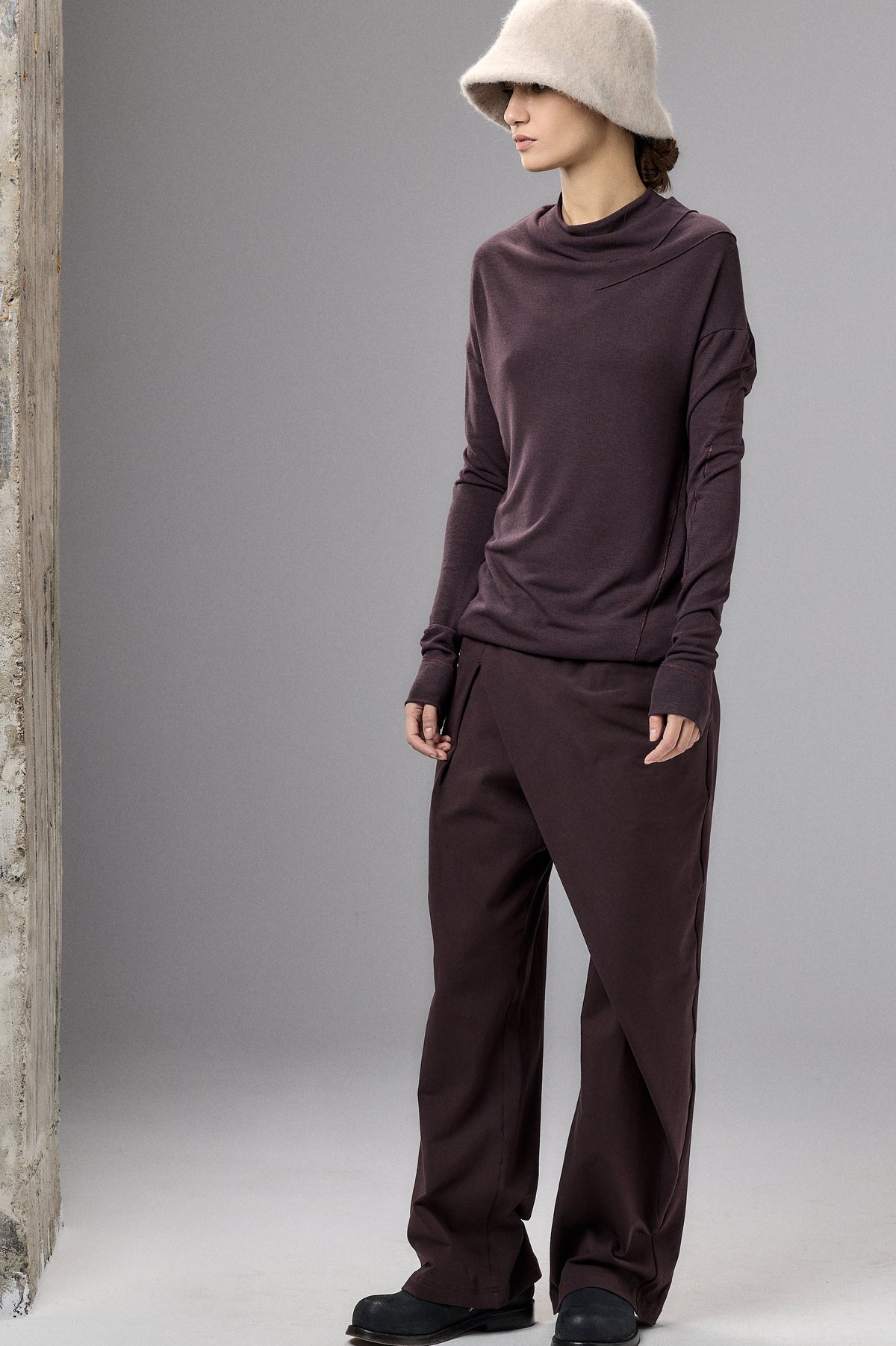 Person wearing a dark plum outfit with a beige hat against a gray background