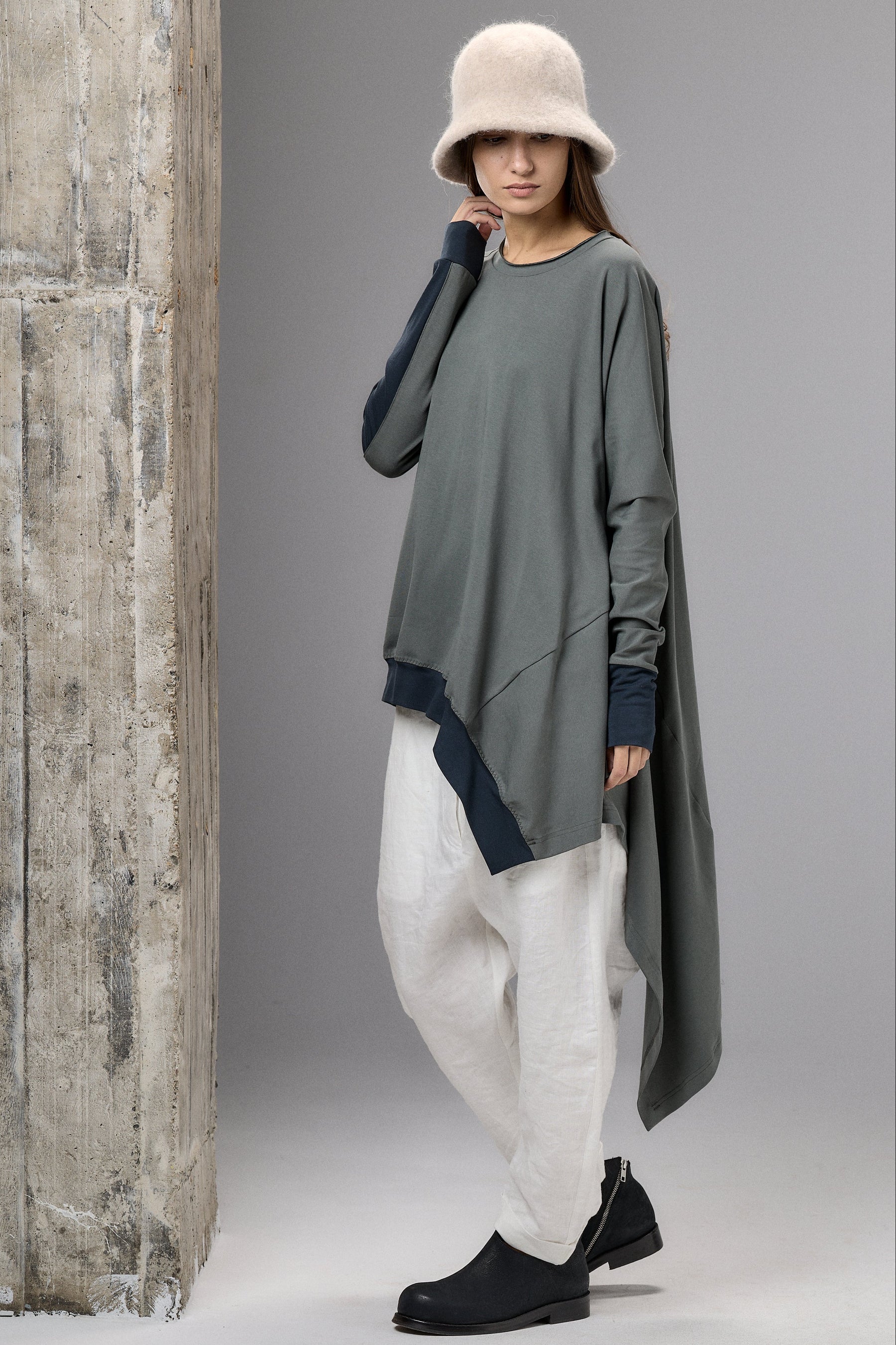 Person wearing a long, dark green asymmetric dress with white pants and a beige hat against a gray background.