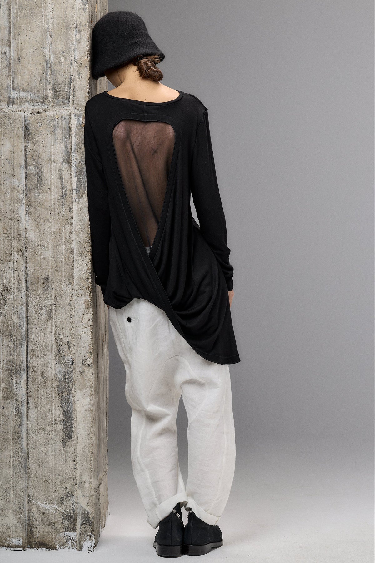 Person wearing a black long-sleeve top with sheer back and white pants against a gray wall.
