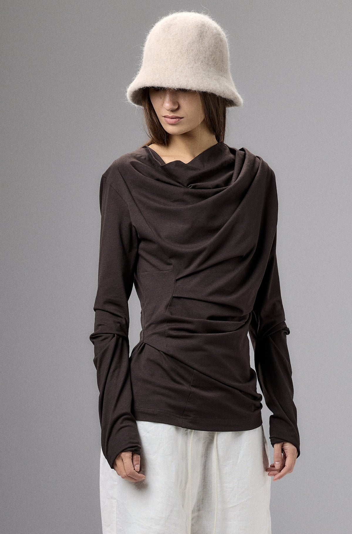 Person wearing a brown long-sleeve top, white pants, and a beige bucket hat against a gray background.