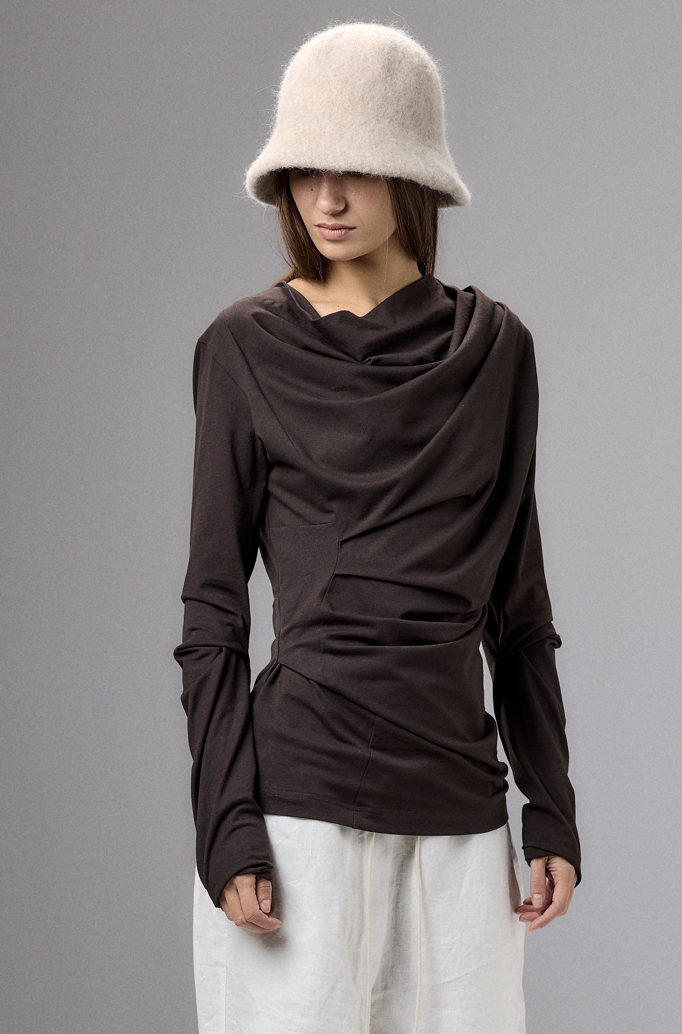 Person wearing a brown long-sleeve top, white pants, and a beige bucket hat against a gray background.