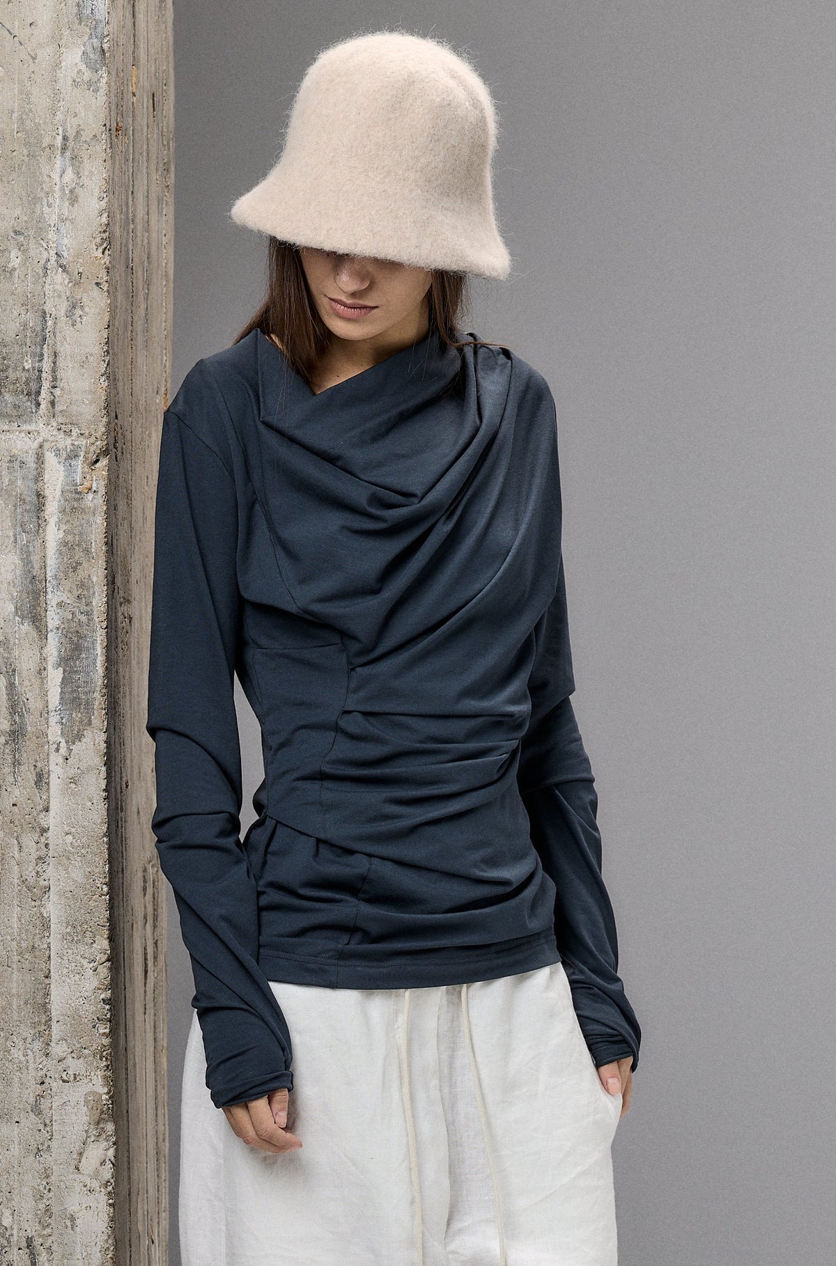 Person wearing a dark long-sleeve top, white pants, and a beige hat against a textured wall.