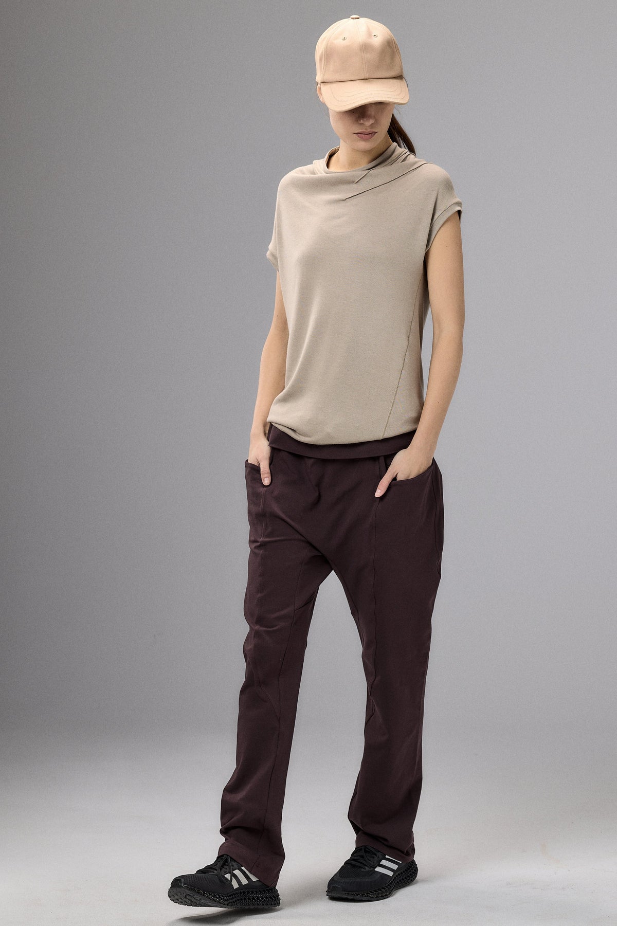 Person wearing a beige sleeveless top and brown pants with a beige cap on a gray background