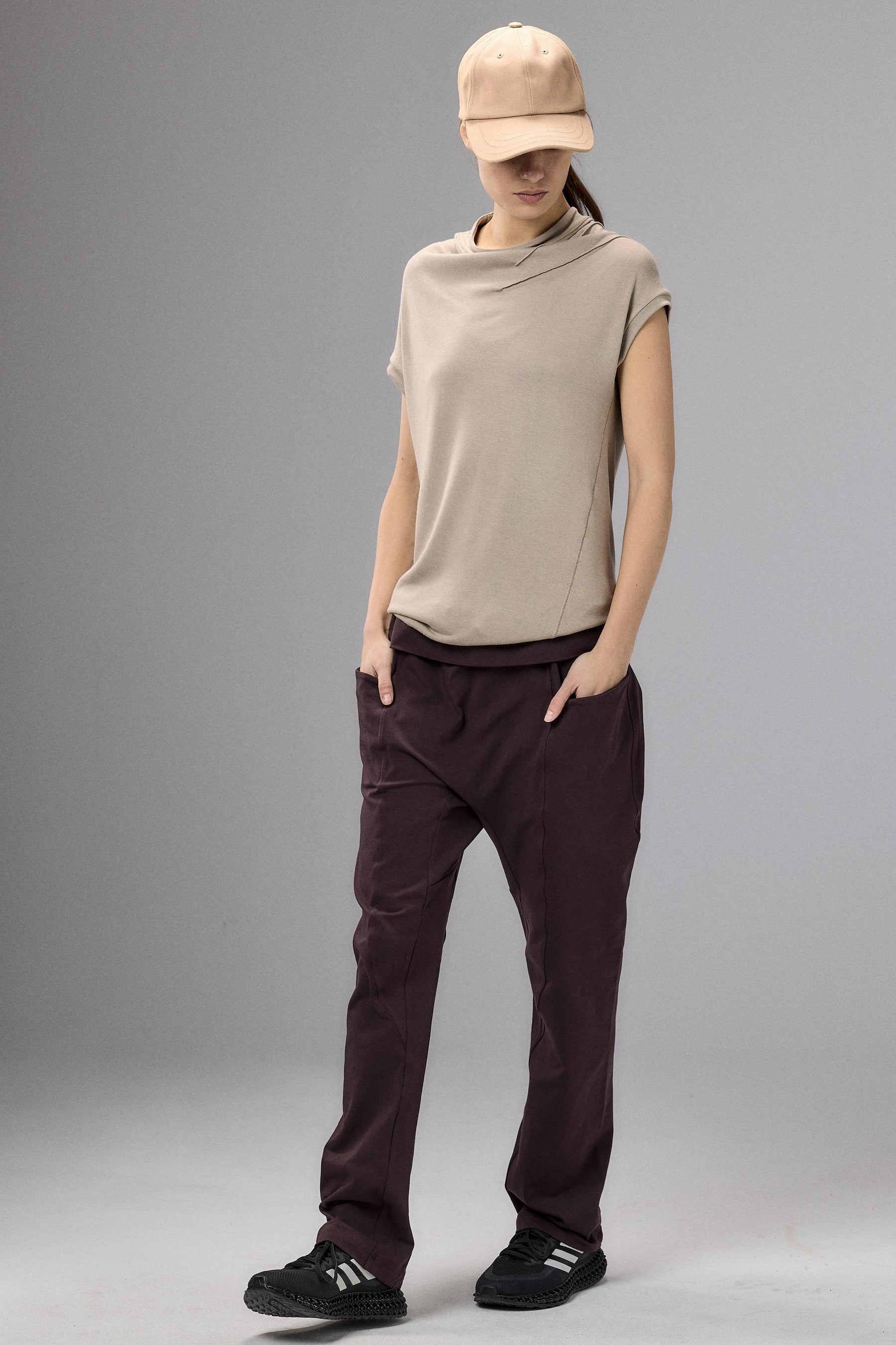 Person wearing a beige sleeveless top and brown pants with a beige cap on a gray background