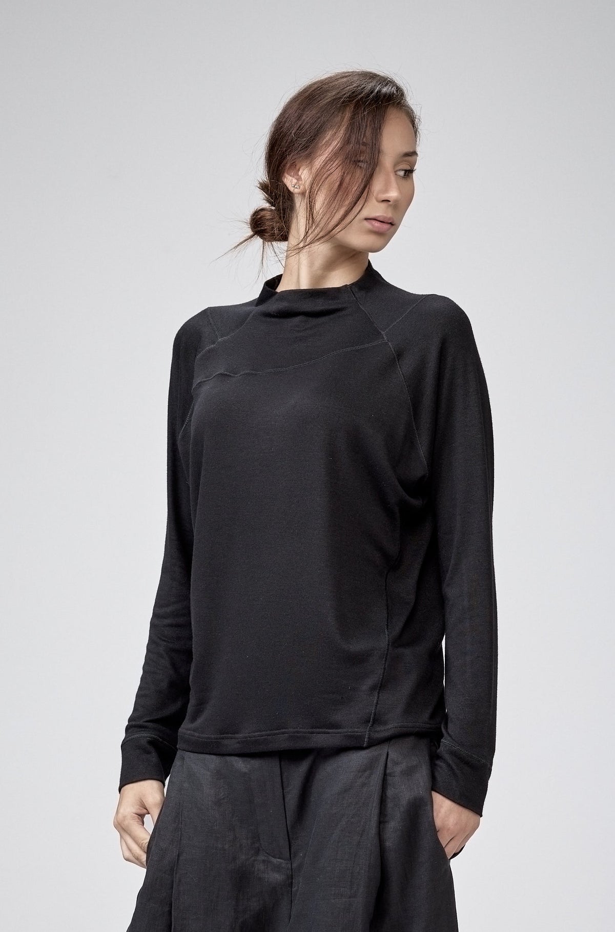 A model wearing a black long-sleeve mock-neck top and matching black baggy pants, standing against a light gray studio background.