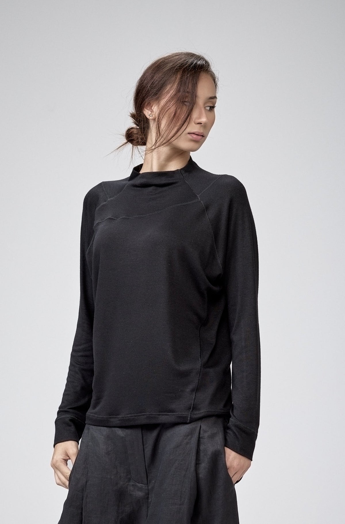 A model wearing a black long-sleeve mock-neck top and matching black baggy pants, standing against a light gray studio background.