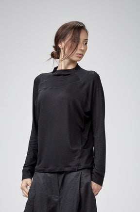 A model wearing a black long-sleeve mock-neck top and matching black baggy pants, standing against a light gray studio background.