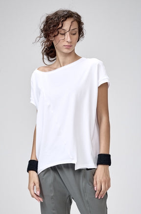 A woman modeling a white off-the-shoulder short-sleeve top and dark gray jogger pants with black cuffs, standing barefoot against a light gray studio background.